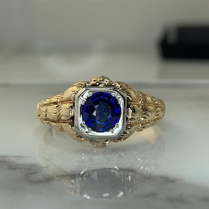 May include: A gold ring with a blue sapphire gemstone set in a white metal bezel. The ring band is intricately designed with a textured pattern.