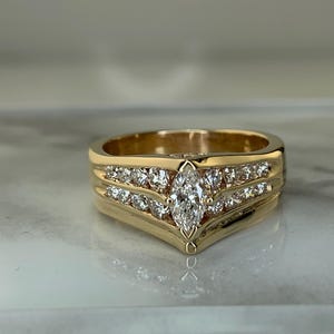 May include: A gold ring with a marquise-cut diamond center stone and smaller round diamonds. The ring has a V-shaped design and is set with multiple rows of sparkling stones, creating a luxurious look. The ring is displayed on a marble surface.