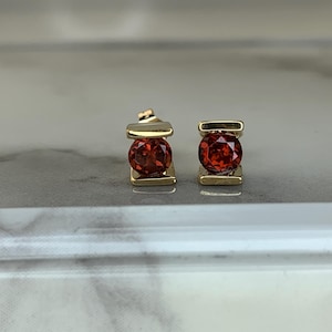 May include: A pair of gold-colored stud earrings, each featuring a vibrant, round, red gemstone. The gemstones are set within a rectangular gold frame, creating a classic and elegant design. The earrings are displayed on a light-colored surface.