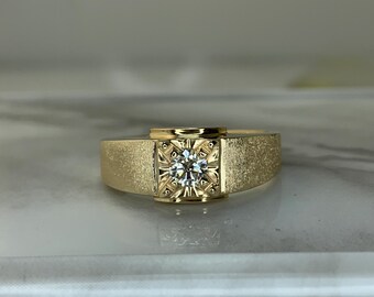 May include: A gold ring with a square, textured center setting. The center holds a round, clear gemstone. The band has a textured finish. The ring is set against a neutral background.