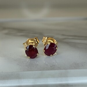 May include: A pair of gold stud earrings, each featuring a vibrant, oval-cut ruby gemstone. The earrings are set in a classic, minimalist design, showcasing the rich red color of the rubies. The studs are displayed on a light-colored surface.