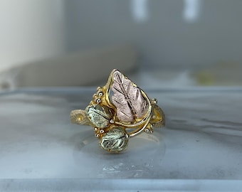 May include: A gold ring with a leaf design, featuring three leaves and a textured band. The leaves are detailed with gold, green, and rose gold tones. The ring is set against a blurred background.