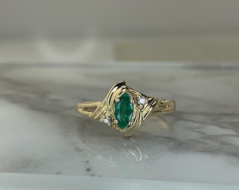 May include: A gold ring with a marquise-shaped emerald green gemstone and two smaller round diamonds. The ring has a delicate, swirling design.