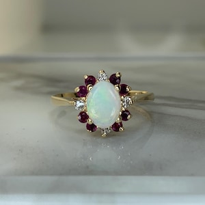 May include: A gold ring featuring a central oval-shaped opal gemstone surrounded by a cluster of small, round, deep red rubies and tiny clear diamonds. The stones are set in a yellow gold band with a polished finish and a classic prong setting.