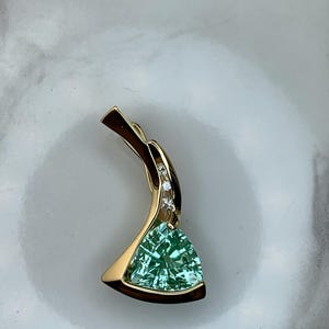 May include: A gold pendant featuring a triangular, faceted, light green gemstone. The pendant has a curved design with small, clear accent stones and a polished finish. The gemstone is approximately 0.75 inches across.