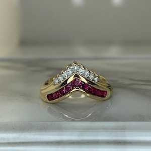 May include: A gold ring with a chevron design. The ring features a row of small, round, red gemstones and a row of small, round, clear gemstones. The ring is set on a white surface.