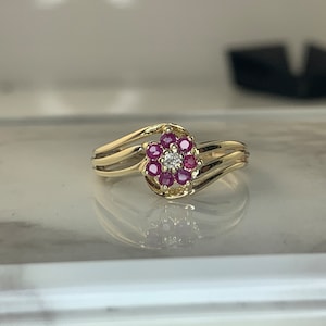 May include: A gold ring with a flower-shaped design featuring a cluster of red rubies and a single white diamond in the centre.