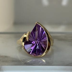 May include: A gold ring featuring a large, faceted, teardrop-shaped purple gemstone. The gemstone is set in a gold band with a unique, asymmetrical design. The ring is displayed on a reflective surface.