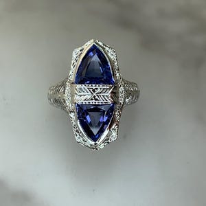 May include: A silver-toned ring featuring two triangular-cut deep blue stones stacked vertically. The center is divided by a horizontal metal band with engraved cross-hatching. The ring band and setting display intricate, openwork filigree metal detailing.