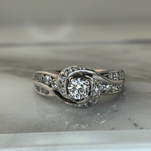 May include: A silver engagement ring with a round center diamond surrounded by smaller diamonds. The band is twisted and features a row of diamonds on each side.