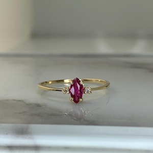 May include: A delicate gold ring featuring a marquise-cut ruby gemstone as the centerpiece. The ring is accented with two small, round diamonds on either side of the ruby. The band is thin and polished, reflecting the light.
