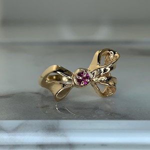 May include: A gold-colored ring shaped like a bow, with a small, round, red gemstone at the center. The ring has a polished finish and is set against a light-colored background.