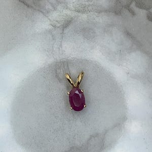 May include: A gold pendant featuring an oval-cut ruby gemstone. The ruby is a deep red color and is set in a gold bezel. The pendant has a simple, elegant design.