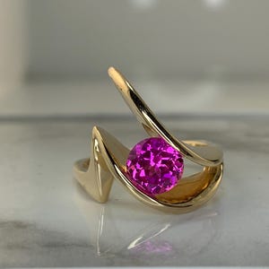 May include: A gold-colored ring with a vibrant pink gemstone. The ring features a unique, abstract design with curved, flowing lines that cradle the central stone. The bright pink color of the gemstone contrasts with the gold.