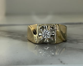 May include: A gold ring with a single round diamond set in a square-shaped bezel. The ring has a textured band.