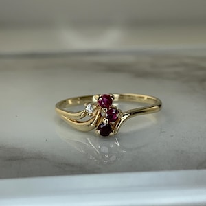 May include: A gold ring with three red gemstones and a single diamond. The ring has a flowing design. The gemstones are round and set in a gold band. The ring is a piece of jewellery.