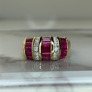 May include: A gold ring featuring a central row of baguette-cut red rubies flanked by two vertical channels of round brilliant-cut white diamonds, with additional baguette-cut rubies set on the outer sides of the band in a wide, multi-row design.