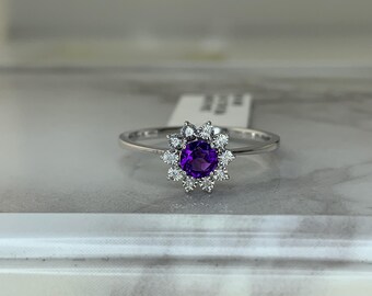 May include: A silver ring with a central, round, deep purple gemstone encircled by smaller, clear gemstones. The ring's design is floral, with the purple stone at the core. The band is slender and polished.