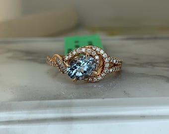 May include: Rose gold ring featuring a light blue oval gemstone, surrounded by a halo of small, clear gemstones. The band has a split design with additional small gemstones. The ring is set on a white surface.