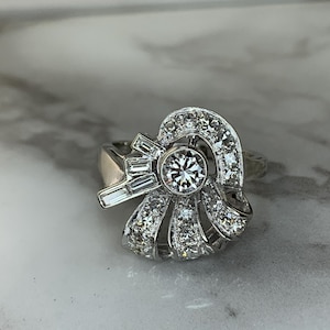 May include: A silver ring with a central round diamond and surrounding smaller diamonds. The ring has a unique, stylized design with baguette-cut diamonds and a fan-like arrangement of stones, set against a marble surface.