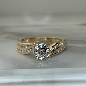 May include: A gold ring with a large, round, clear gemstone as the centerpiece. The band features smaller, clear gemstones set in rows on either side of the main stone. The ring is displayed on a light-coloured surface.