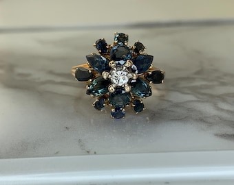 May include: A gold ring featuring a floral design with dark blue gemstones and a central white gemstone. The ring's design is reminiscent of a flower, with the gemstones arranged to create petals.