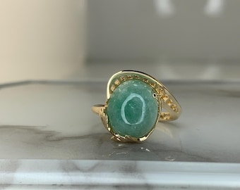 May include: A gold ring featuring a large, oval-shaped, light green gemstone. The ring has a decorative gold band that curves around the gemstone. The ring is set on a white surface.