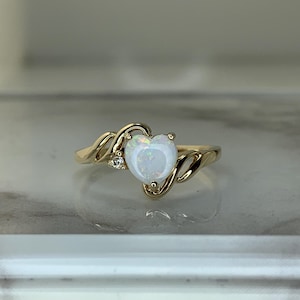 May include: A gold ring featuring a heart-shaped opal gemstone. The opal is white with iridescent qualities, set in a swirling gold band. A small diamond accent is set on the side of the heart. Jewelry.