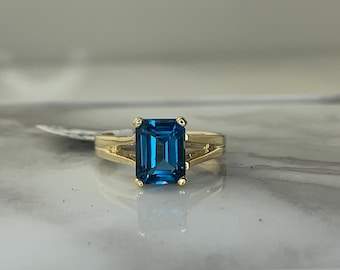 May include: A gold ring featuring a blue topaz gemstone. The ring has a split shank design.