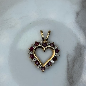 May include: A gold heart pendant, featuring a halo of red gemstones and small white stones. The pendant has a heart-shaped void in the centre and two small loops at the top for a chain. The gemstones are evenly spaced around the heart.
