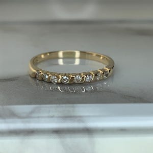 May include: A gold band ring featuring a series of small, round, clear stones set in a channel along the front half of the band. The metal has a polished, reflective finish, and the stones are arranged in a continuous row across the top section.