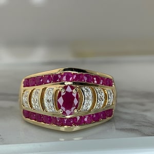 May include: A gold ring featuring a central oval-cut ruby surrounded by two rows of smaller round rubies. Between the ruby rows are two curved sections set with small white diamonds. The band is polished gold with a wide, tapered design.