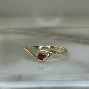 May include: A gold ring with a flowing design. The ring features a round, deep red gemstone and a small, clear gemstone. The ring is set on a light-coloured surface.