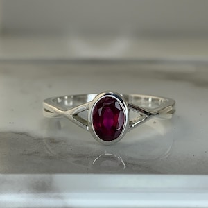 May include: A silver ring featuring an oval, deep red gemstone. The band has a unique, interwoven design. The ring is displayed on a reflective surface, highlighting its polished finish. A piece of jewelry.
