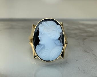 May include: A gold ring featuring an oval cameo with a profile of a woman. The cameo has a light blue figure against a black background. The ring has a gold band with decorative elements on the sides.