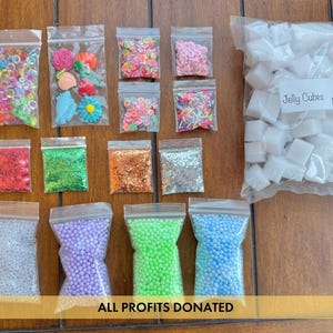 May include: A collection of craft supplies in clear plastic bags, including colorful beads, glitter, and decorative shapes. A large bag of white jelly cubes is also present. The text "ALL PROFITS DONATED" is visible.