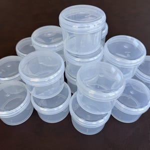 May include: A collection of clear, round plastic containers with lids. The containers are stacked and scattered on a dark brown surface. The containers are made of a translucent plastic material.
