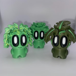May include: Three green figurines with plant-like features. Each has large, oval eyes and a small smile. The figurines have different leafy or floral headpieces. The background is plain white.
