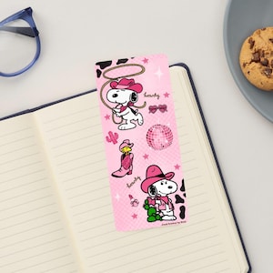 May include: A pink bookmark featuring Snoopy in cowboy attire with the word "howdy". The bookmark has a cowhide pattern at the top and includes a disco ball and cactus graphic. The bookmark is on a notebook.