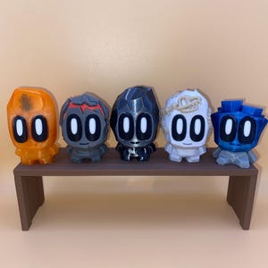 May include: Five colorful, stylized figurines with large oval eyes and geometric bodies are displayed on a brown shelf. The figurines are orange, gray, black, white, and blue. The shelf is brown and has a simple, rectangular design.