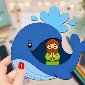May include: A layered paper craft cutout featuring a dark blue whale with a light blue underbelly and a water spout. Inside the whale's open mouth is a cartoon illustration of a person with brown hair and a beard, wearing a green tunic.
