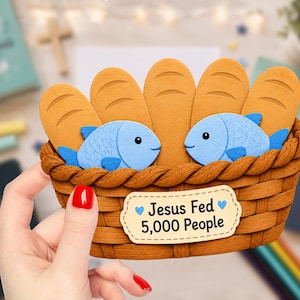 May include: A layered paper craft depicting a brown woven basket filled with four tan bread loaves and two light blue fish. A rectangular label on the front of the basket features the text "Jesus Fed 5,000 People" with two small blue hearts.