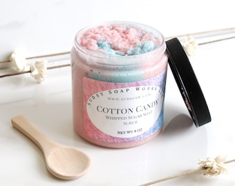 May include: A jar of cotton candy whipped sugar soap with a wooden spoon. The soap is pink and blue, and the jar has a black lid. The label reads "Cotton Candy Whipped Sugar Soap Scrub" and "Net Wt. 8 oz."