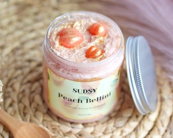 May include: A jar of peach-scented sugar scrub with a light pink hue and gold flecks. The jar is labeled "SUDSY Peach Bellini" and contains three peach-colored soap pieces. The lid is silver.