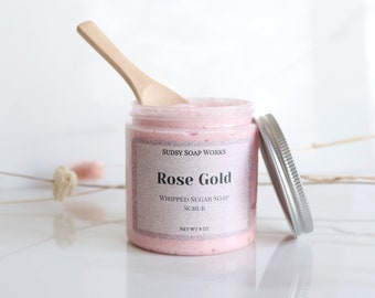 Może przedstawiać: Różowy peeling do ciała z mydła cukrowego w słoiku szklanym z srebrną pokrywką. Słoik ma etykietę z napisem "Sudsy Soap Works" i "Rose Gold Whipped Sugar Soap Scrub" o wadze netto 8 uncji.