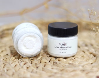 May include: Two small, clear jars of lip scrub on a woven mat. One jar is open, revealing a white, textured scrub. The other jar has a black lid and a label that reads "SUDSY Marshmallow LIP SCRUB Net Wt. 28g."
