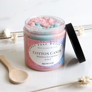 May include: A clear jar of cotton candy whipped sugar soap scrub with a black lid and a small wooden spoon. The jar has a pink, blue, and white swirl design. The label reads "Cotton Candy" and "Whipped Sugar Soap Scrub".