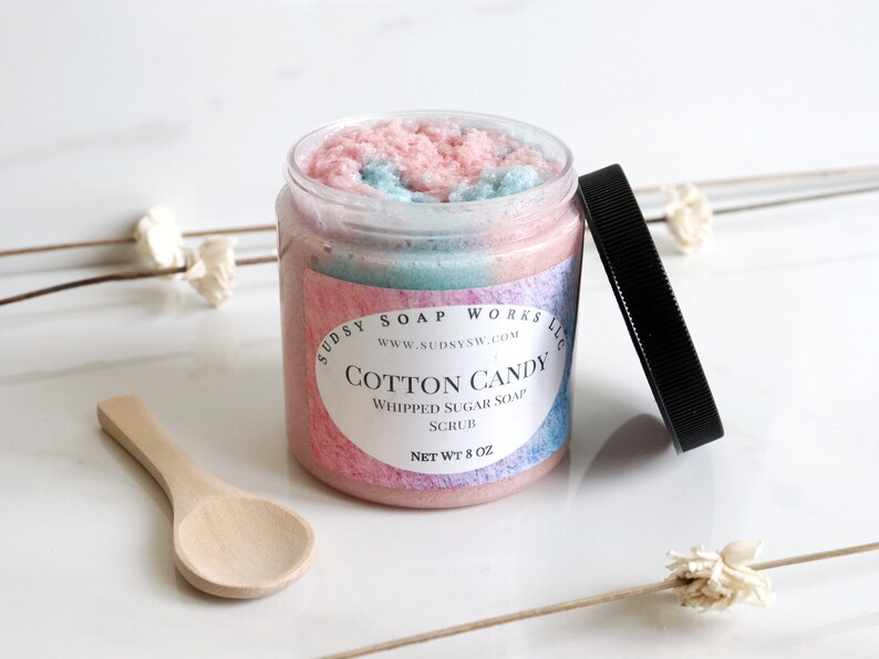 May include: A clear jar of cotton candy whipped sugar soap scrub with a black lid and a small wooden spoon. The jar has a pink, blue, and white swirl design. The label reads "Cotton Candy" and "Whipped Sugar Soap Scrub".