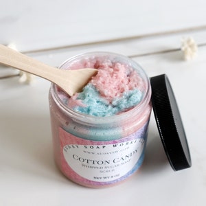 May include: A jar of cotton candy whipped sugar soap scrub with a wooden spoon. The scrub is pink and blue, and the jar has a black lid. The label says "Cotton Candy" and "Whipped Sugar Soap Scrub".