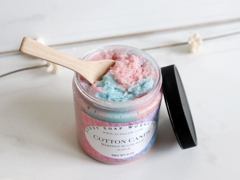 May include: A jar of cotton candy whipped sugar soap scrub with a wooden spoon. The scrub is pink and blue, and the jar has a black lid. The label says "Cotton Candy" and "Whipped Sugar Soap Scrub".
